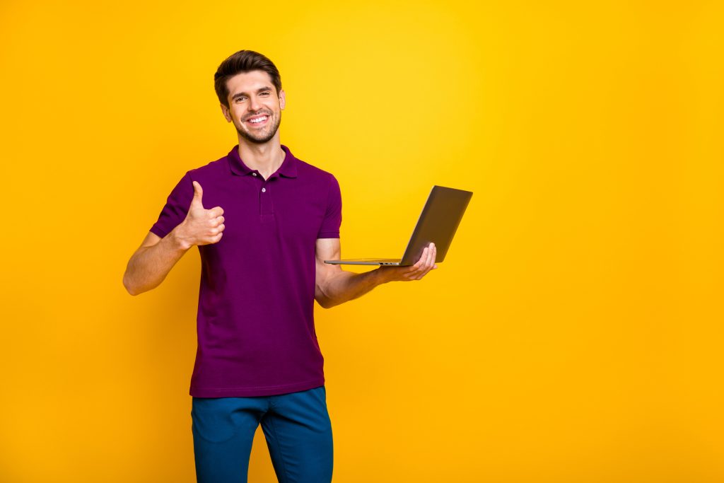 Photo of cheerful positive nice attractive man showing you thumb up, holding laptop wearing blue pants trousers isolated over yellow vivid color background