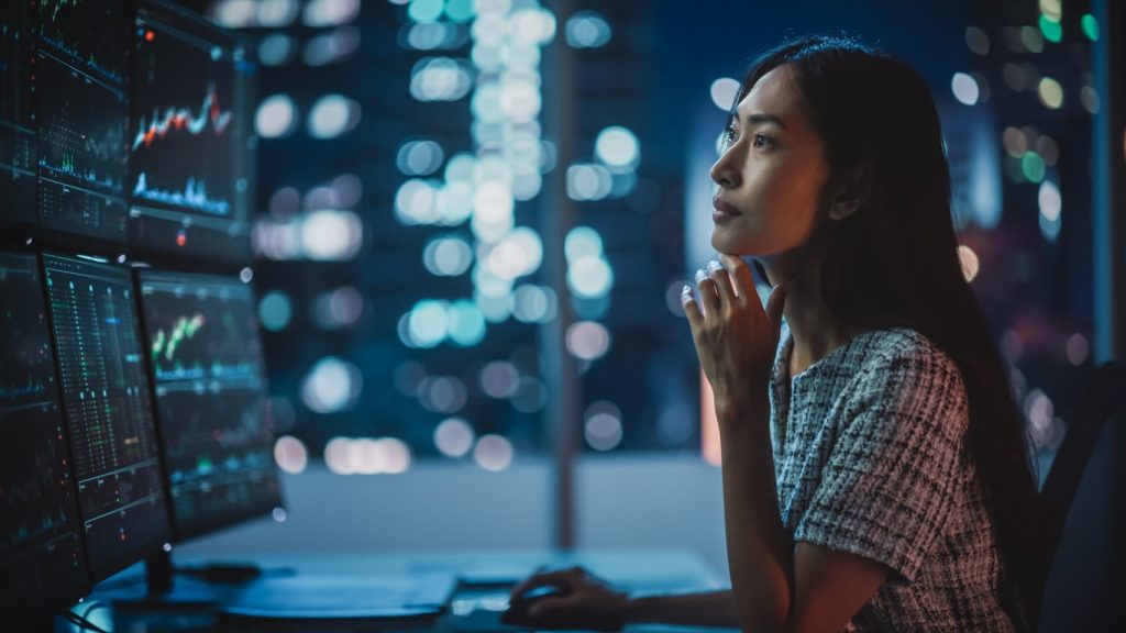 Portrait of a Financial Analyst Working on Computer with Multi-Monitor Workstation with Real-Time Stocks, Commodities and Exchange Market Charts. Businesswoman at Work in Investment Broker Agency.